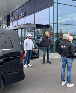 Vier Männer stehen vor der modernen Glasfassade des Mercedes-Benz Autohauses Peter. Links ragt das Heck einer schwarzen V-Klasse ins Bild.