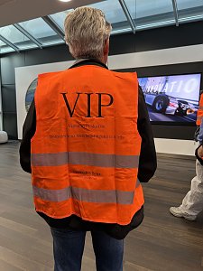 Ein Mann von hinten gesehen trägt eine leuchtend orangefarbene Warnweste mit der großen Aufschrift VIP. Im Hintergrund ist ein Bildschirm zu erkennen, auf dem ein Lkw abgebildet ist.