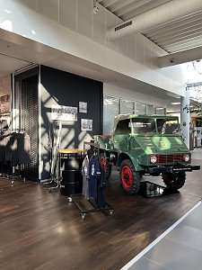 Ein historischer, grüner Unimog mit roten Felgen steht als Ausstellungsstück auf einem dunklen Holzboden. Links daneben sind Kleiderständer mit Merchandise-Artikeln und dekorative schwarze Fässer platziert.